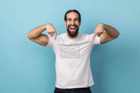Foto de Look at ads below. Portrait of man with beard wearing white T-shirt smiling and pointing down, showing place for idea presentation, commercial text. Indoor studio shot isolated on blue background. - Imagen libre de derechos