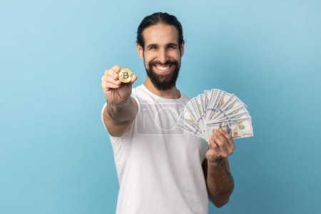 Photo for Portrait of happy man with beard wearing white T-shirt showing bitcoin and big fan of dollars banknotes, e-commerce, crypto currency. Indoor studio shot isolated on blue background. - Royalty Free Image