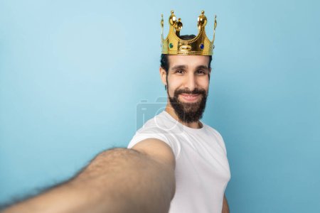 Téléchargez les photos : Portrait of satisfied handsome man with beard wearing white T-shirt and gold crown, looking at camera with smile, POV, point of view of photo. Indoor studio shot isolated on blue background. - en image libre de droit