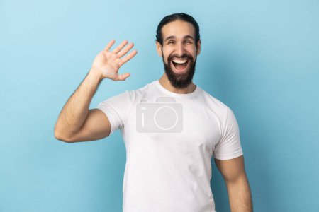 Photo for Portrait of positive man with beard wearing white T-shirt standing with raised palm gesturing hi to camera, welcoming with toothy smile. Indoor studio shot isolated on blue background. - Royalty Free Image