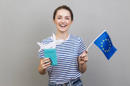 Téléchargez les photos : Portrait of woman wearing striped T-shirt standing with Europe union flag and passport with paper plane, traveling to european country. Indoor studio shot isolated on gray background. - en image libre de droit