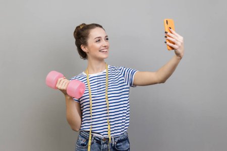 Foto de Portrait of woman wearing striped T-shirt making selfie, looking at camera of smartphone, pumping up muscles and working out at gym. Indoor studio shot isolated on gray background. - Imagen libre de derechos