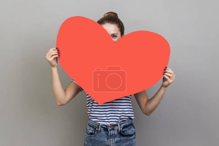 Téléchargez les photos : Portrait of woman hiding face behind big red heart and looking at camera with curious prying eyes, expressing i love you, feeling affection fondness. Indoor studio shot isolated on gray background. - en image libre de droit