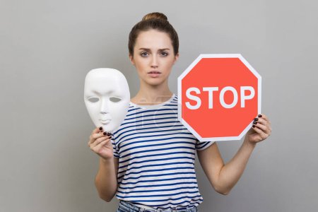 Photo for Portrait of serious woman wearing striped T-shirt holding white mask with unknown face and red traffic sign, looking at camera. Indoor studio shot isolated on gray background. - Royalty Free Image