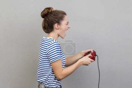 Foto de Side view portrait of woman wearing striped T-shirt holding gamepad, winning video games competition, screaming happy expression. Indoor studio shot isolated on gray background. - Imagen libre de derechos