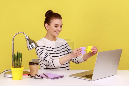 Photo for Satisfied woman showing to laptop display two colored pieces of jigsaw, easily solving difficult tasks at work, sitting at workplace. Indoor studio studio shot isolated on yellow background. - Royalty Free Image