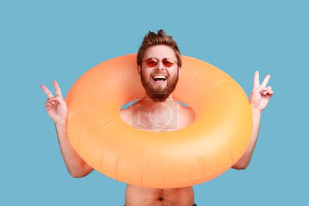 Téléchargez les photos : Portrait of excited handsome bearded man holding bright orange rubber ring, looking at camera, enjoying his vacation, showing v sign. Indoor studio shot isolated on blue background. - en image libre de droit