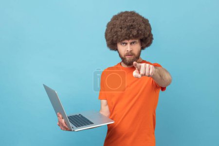 Téléchargez les photos : Strict bossy man with Afro hairstyle wearing orange T-shirt holding laptop in hands, working online, pointing finger to camera with serious expression. Indoor studio shot isolated on blue background. - en image libre de droit