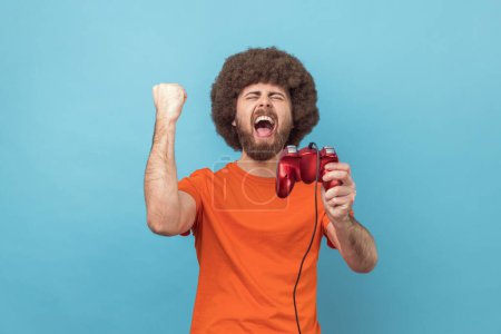 Téléchargez les photos : Man with Afro hairstyle wearing orange T-shirt holding hand red joystick pad showing yes gesture sincerely rejoicing, victory in online virtual game. Indoor studio shot isolated on blue background. - en image libre de droit