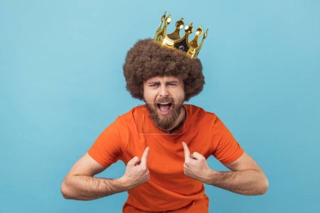 Foto de Portrait of crazy man with Afro hairstyle in golden crown and pointing himself with excite expression, looking with arrogance, declaring his authority. Indoor studio shot isolated on blue background. - Imagen libre de derechos