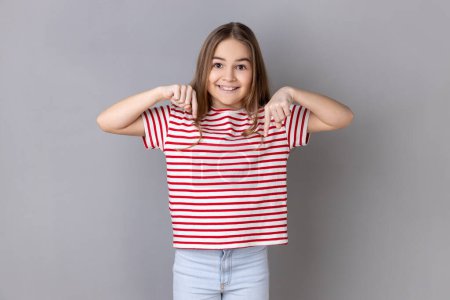 Foto de Look, advertise below. Portrait of little girl wearing striped T-shirt pointing down place for commercial idea, looking at camera with toothy smile. Indoor studio shot isolated on gray background. - Imagen libre de derechos