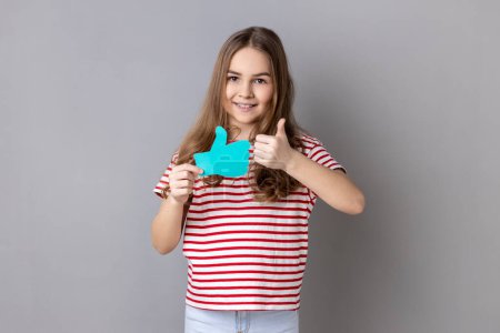 Foto de Little girl wearing striped T-shirt holding thumbs up blue icon, follower notification symbol, recommendation and good feedback in social media. Indoor studio shot isolated on gray background. - Imagen libre de derechos