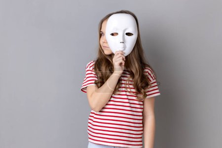 Photo for Portrait of dark haired little cute girl wearing striped T-shirt holding white mask and looking away with serious facial expression. Indoor studio shot isolated on gray background. - Royalty Free Image