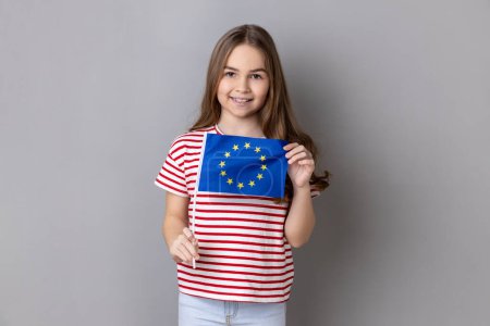 Foto de European Union flag. Portrait of smiling adorable cute little girl wearing striped T-shirt holding Europe flag, looking at camera. Indoor studio shot isolated on gray background. - Imagen libre de derechos