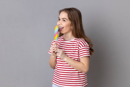 Foto de Portrait of little girl wearing striped T-shirt holding colorful candy, licking, looking away with childish satisfied expression. Indoor studio shot isolated on gray background. - Imagen libre de derechos