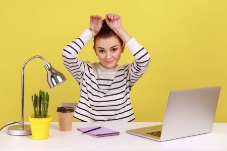 Foto de Charming woman manager smiling and showing bunny ears on head, resting, having fun at workplace in home office, feeling carefree and happy. Indoor studio studio shot isolated on yellow background. - Imagen libre de derechos