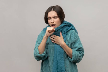 Portrait of woman wrapped in warm scarf, coughing, feeling unwell suffering fever, seasonal influenza symptoms, wearing casual style jacket. Indoor studio shot isolated on gray background.