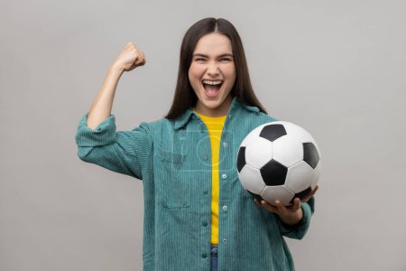 Téléchargez les photos : Satisfied woman screaming widely opening mouth, celebrating victory of favourite football team, holding ball in hands, wearing casual style jacket. Indoor studio shot isolated on gray background. - en image libre de droit