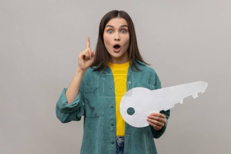 Foto de Surprised woman pointing finger up having idea and holding big paper key, looking amazed by sudden solution, wearing casual style jacket. Indoor studio shot isolated on gray background. - Imagen libre de derechos