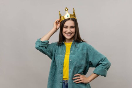 Téléchargez les photos : Woman in crown on head looking at with confident expression, self-motivation and dreams to be best. keeping hand on hip, wearing casual style jacket. Indoor studio shot isolated on gray background. - en image libre de droit