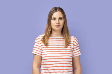 Foto de Portrait of serious confident blond woman wearing striped T-shirt standing looking at camera with attentive look, being strict and bossy. Indoor studio shot isolated on purple background. - Imagen libre de derechos