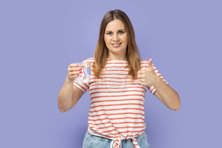Foto de Portrait of blond woman wearing striped T-shirt holding glass of water, showing thumb up, like fresh beverage, having happy expression. Indoor studio shot isolated on purple background. - Imagen libre de derechos