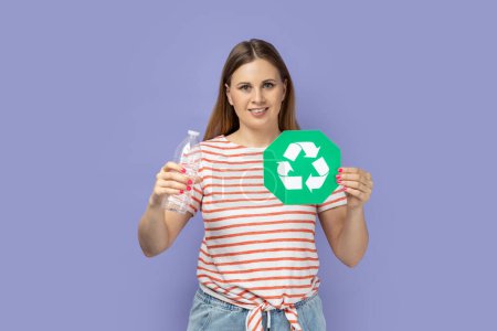 Foto de Portrait of pretty optimistic good looking blond woman wearing striped T-shirt showing green recycling sign and holding plastic bottle in hands. Indoor studio shot isolated on purple background. - Imagen libre de derechos