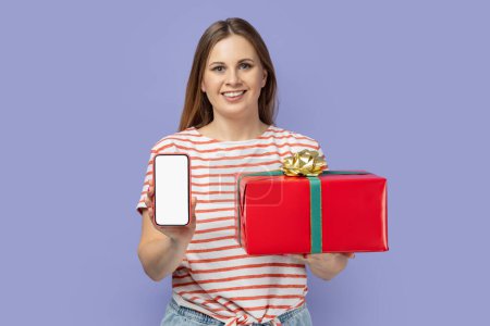 Photo for Portrait of satisfied blond woman wearing striped T-shirt holding red present box and showing smart phone with white empty display. Indoor studio shot isolated on purple background. - Royalty Free Image