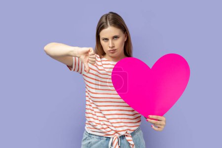 Téléchargez les photos : Portrait of sad upset blond woman wearing striped T-shirt holding big pink heart , looking at camera, expressing sadness, showing thumb down gesture. Indoor studio shot isolated on purple background. - en image libre de droit