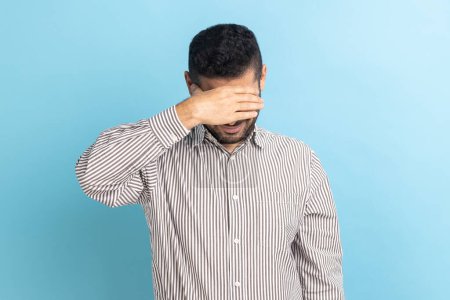 Photo for Young adult businessman closing eyes with hand, dont want to see that, ignoring problems, hiding from stressful situations, wearing striped shirt. Indoor studio shot isolated on blue background. - Royalty Free Image