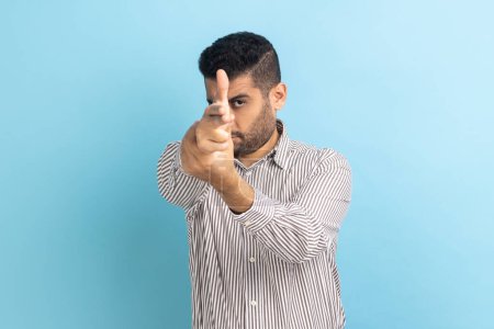 Téléchargez les photos : Portrait of dangerous young adult handsome businessman with beard pointing with finger pistols at camera, pretending to shoot, wearing striped shirt. Indoor studio shot isolated on blue background. - en image libre de droit