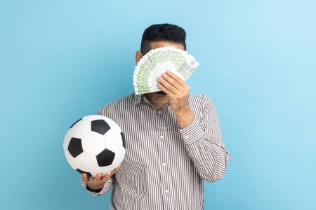 Photo for Unknown anonymous businessman holding soccer ball and covering face with fan of euro bills, sports betting, big win, wearing striped shirt. Indoor studio shot isolated on blue background. - Royalty Free Image