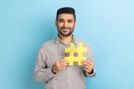 Téléchargez les photos : Viral hashtag and successful blogging. Satisfied businessman with beard holding large yellow hash symbol, wearing striped shirt. Indoor studio shot isolated on blue background. - en image libre de droit
