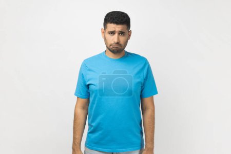 Téléchargez les photos : Portrait of sad upset depressed unshaven man wearing blue T- shirt standing looking at camera with frowning face, expressing sadness, being offended. Indoor studio shot isolated on gray background. - en image libre de droit