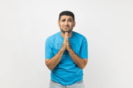 Téléchargez les photos : Portrait of sad upset unshaven man wearing blue T- shirt standing has sorrowful expression asking somebody to forgive, keeps palms in praying gesture. Indoor studio shot isolated on gray background. - en image libre de droit