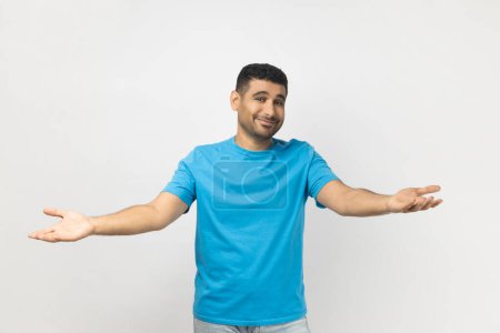 Photo for Portrait of unshaven man wearing blue T- shirt standing shows welcome gesture, spreads hands as wants to cuddle best friend, smiles broadly. Indoor studio shot isolated on gray background. - Royalty Free Image