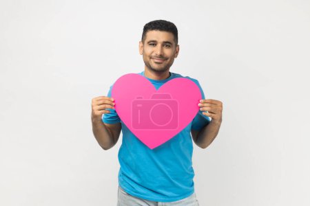 Photo for Portrait of romantic smiling joyful unshaven man wearing blue T- shirt standing holding big pink heart, expressing fondness and love. Indoor studio shot isolated on gray background. - Royalty Free Image