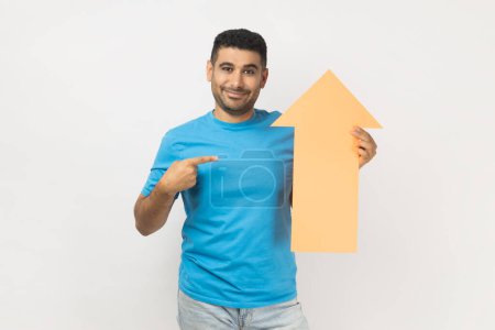 Téléchargez les photos : Portrait of cheerful unshaven man wearing blue T- shirt standing pointing at carton arrow indicating up, being glad of career success. Indoor studio shot isolated on gray background. - en image libre de droit