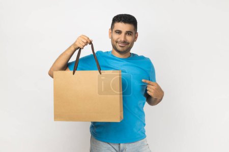 Photo for Portrait of joyful handsome cheerful unshaven man wearing blue T- shirt standing pointing at shopping bag, enjoying spending time in mall. Indoor studio shot isolated on gray background. - Royalty Free Image