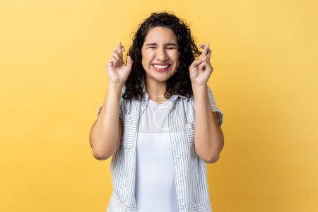Foto de Portrait of hopeful adorable woman with dark wavy hair standing with crossed fingers, wishes her dream comes true, keeps eyes closed. Indoor studio shot isolated on yellow background. - Imagen libre de derechos