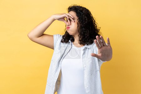 Foto de Portrait of woman with dark wavy hair pinching her nose to hold breath and showing stop gesture, disgusted by unpleasant odor, fart. Indoor studio shot isolated on yellow background. - Imagen libre de derechos