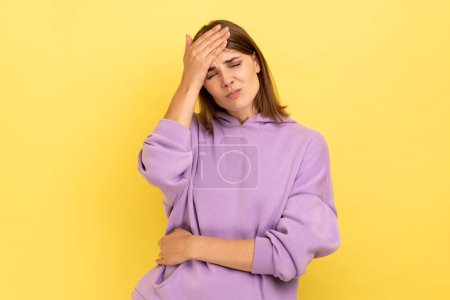 Photo for Sad woman with dark hair making facepalm gesture keeping hand on head, blaming herself for bad memory, unforgivable mistake, wearing purple hoodie. Indoor studio shot isolated on yellow background. - Royalty Free Image