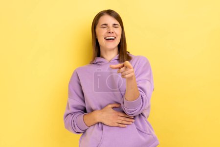 Photo for Hey, you are ridiculous. Woman laughing, holding stomach and pointing to camera, taunting you, can't stop hysterical laughter, wearing purple hoodie. Indoor studio shot isolated on yellow background. - Royalty Free Image