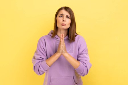 Foto de Portrait of pleading woman keeping arms in prayer gesture and asking forgiveness, feeling sorry for mistake, wearing purple hoodie. Indoor studio shot isolated on yellow background. - Imagen libre de derechos