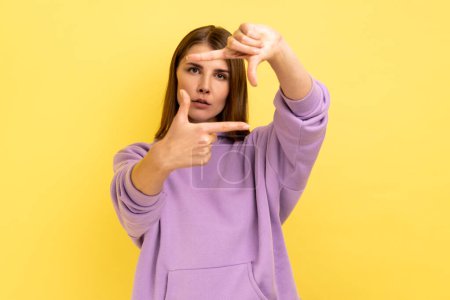 Foto de Serious woman looking attentively through photo frame shape with fingers, making photography gesture, capturing moment, wearing purple hoodie. Indoor studio shot isolated on yellow background. - Imagen libre de derechos