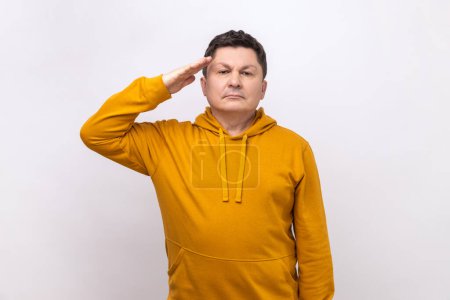 Foto de Subordinate man saluting, holding hand near head, looking at camera with serious expression, patriotism and discipline, wearing urban style hoodie. Indoor studio shot isolated on white background. - Imagen libre de derechos