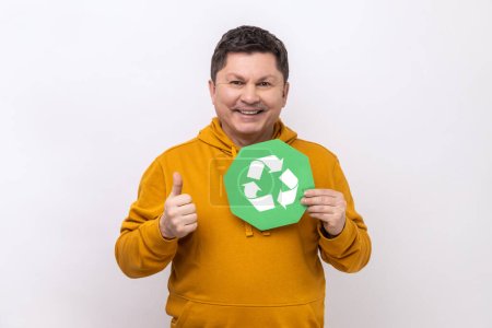 Foto de Man holding green recycling signboard pointing finger at camera, calling for garbage sorting and environment protection, wearing urban style hoodie. Indoor studio shot isolated on white background. - Imagen libre de derechos