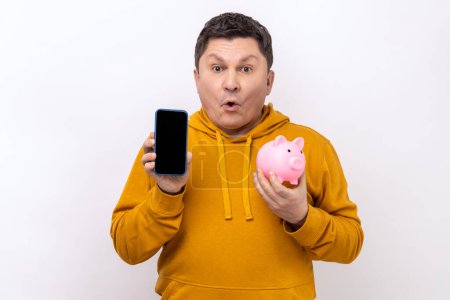 Téléchargez les photos : Astonished shocked man holding smartphone with empty screen and piggy bank, cashback, online wallet, wearing urban style hoodie. Indoor studio shot isolated on white background. - en image libre de droit