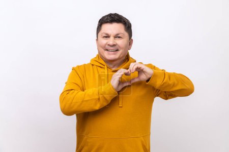 Photo for Happy handsome dark haired man standing with heart or love gesture and looking at camera with toothy smile, wearing urban style hoodie. Indoor studio shot isolated on white background. - Royalty Free Image