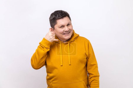 Téléchargez les photos : Portrait of handsome cheerful man holding telephone hand gesture near ear, imitating communication on phone, wearing urban style hoodie. Indoor studio shot isolated on white background. - en image libre de droit